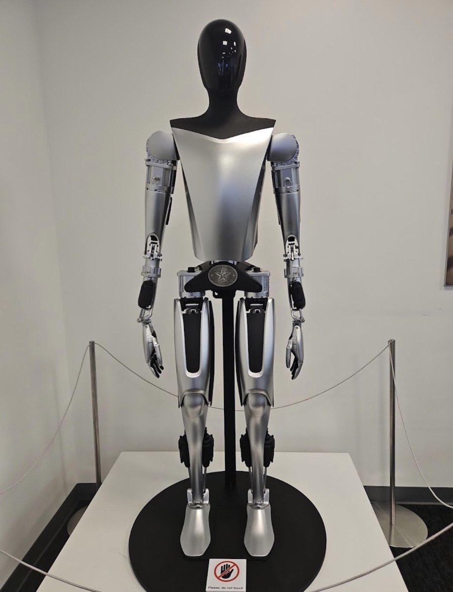 A Tesla Optimus humanoid robot standing on a display platform behind a rope barrier